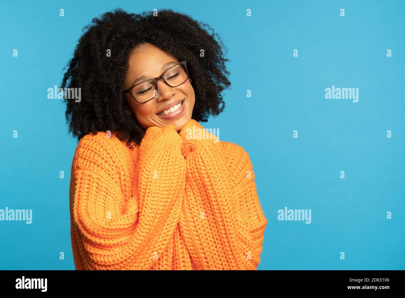 Stylish happy dark skinned millennial woman with curly hair wear orange knitted jumper, holds hands under chin, copy space for advertising, isolated o Stock Photo