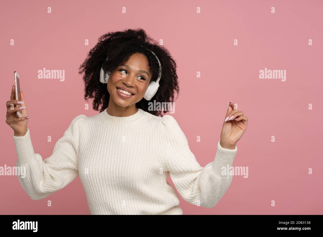 Happy dark skinned millennial woman with headphones enjoying listening to the artist's new album, dances, makes movements to music, holding mobile pho Stock Photo