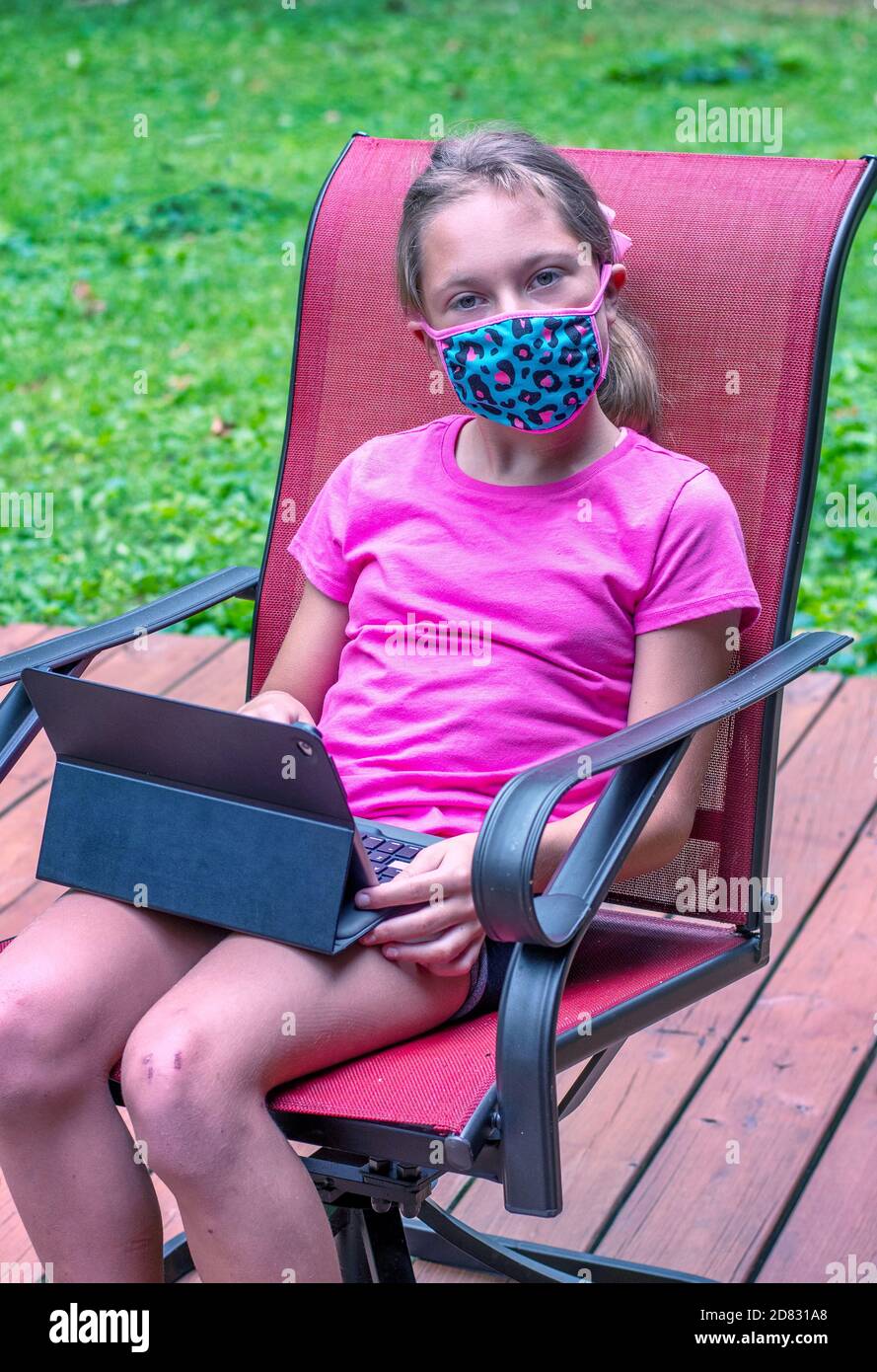 Young girl in a mask works outdoors on a small laptop as she does remote school learning during the Covid-19 pandemic Stock Photo