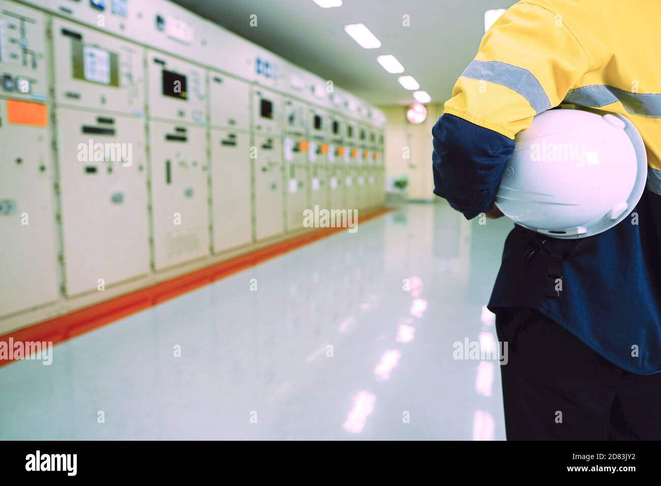 Male engineer wearing a yellow uniform and wearing a white safety hat, inspecting electrical systems in a large power plant. Stock Photo