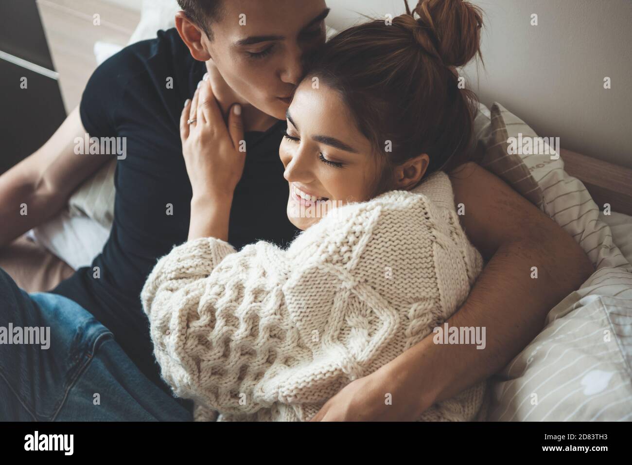 Caucasian man kissing his cute wife dressed in a white knitted sweater embracing in bed Stock Photo