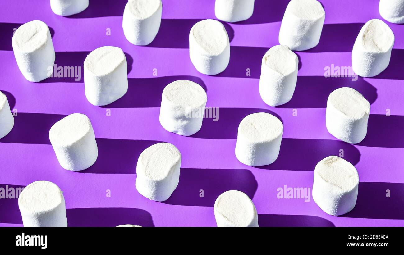 Many rows of white marshmallows cylindrical form lies on purple background. Pattern of unhealthy junky food. Stock Photo