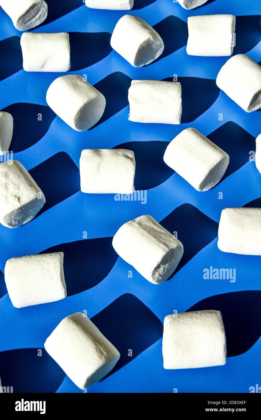 Many rows of white marshmallows cylindrical form lies on blue background. Pattern of unhealthy junky food. Stock Photo