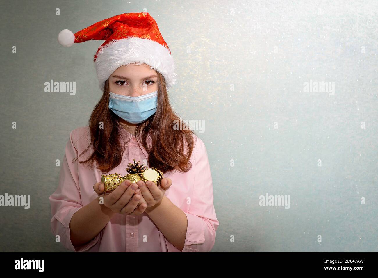 A teenage girl stands at home wearing a Santa hat and a medical mask, holding golden Christmas tree toys in her palms Stock Photo