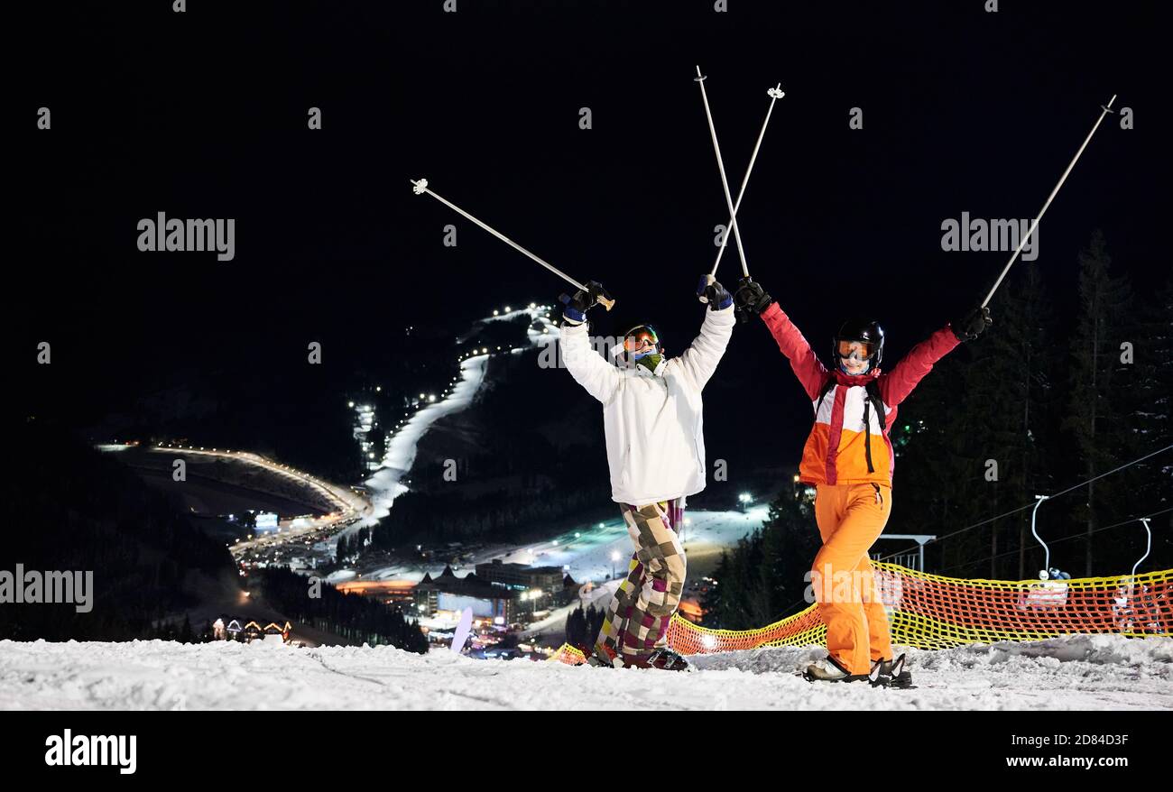 Cheerful male and female skiers in winter jackets and helmets raising ski poles. Joyful young man and lady in ski goggles standing on snow-covered slope with night ski resort on background. Stock Photo