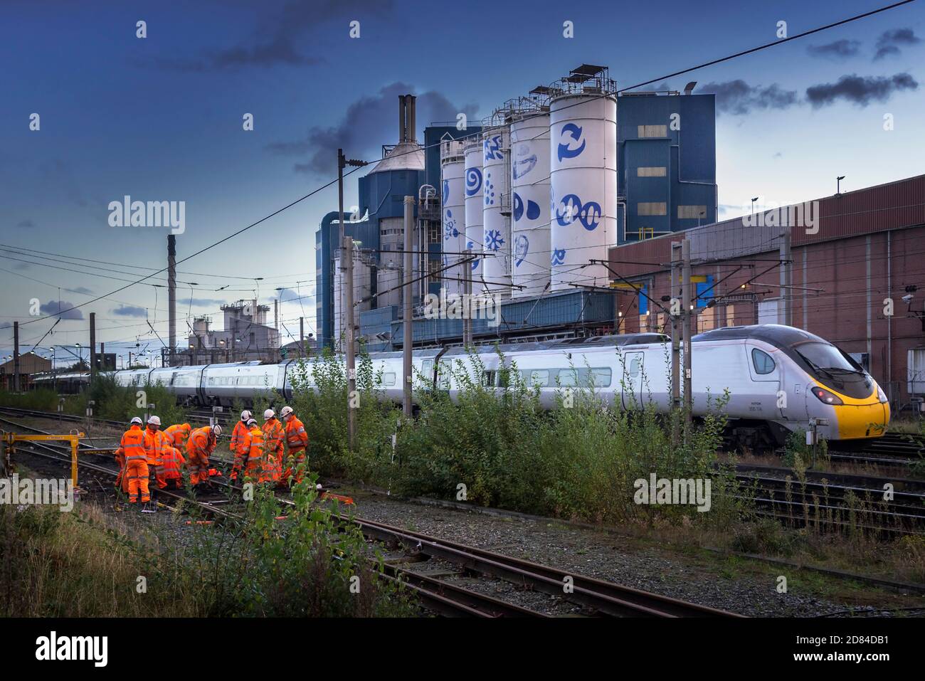 Network Rail track workers busy on the West Coast Main Line at Warrington Bank Quay station with the Unilever soaps factory in the early evening. Stock Photo