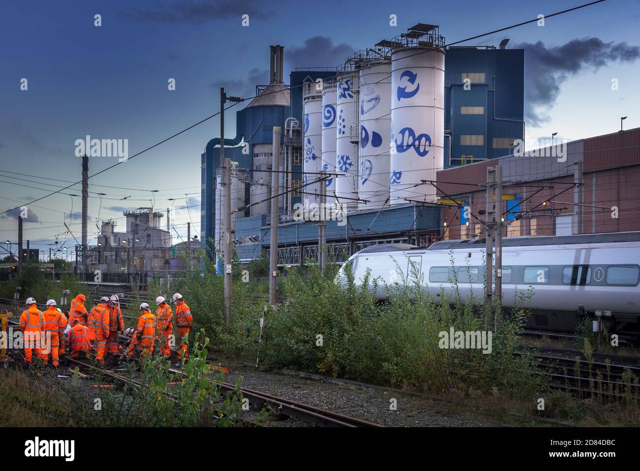 Network Rail track workers busy on the West Coast Main Line at Warrington Bank Quay station with the Unilever soaps factory in the early evening. Stock Photo