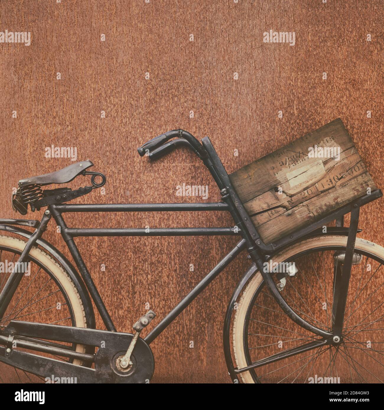 Retro styled image of a black cargo bicycle with old wooden transport crate and leather saddle in front of a corten steel wall Stock Photo