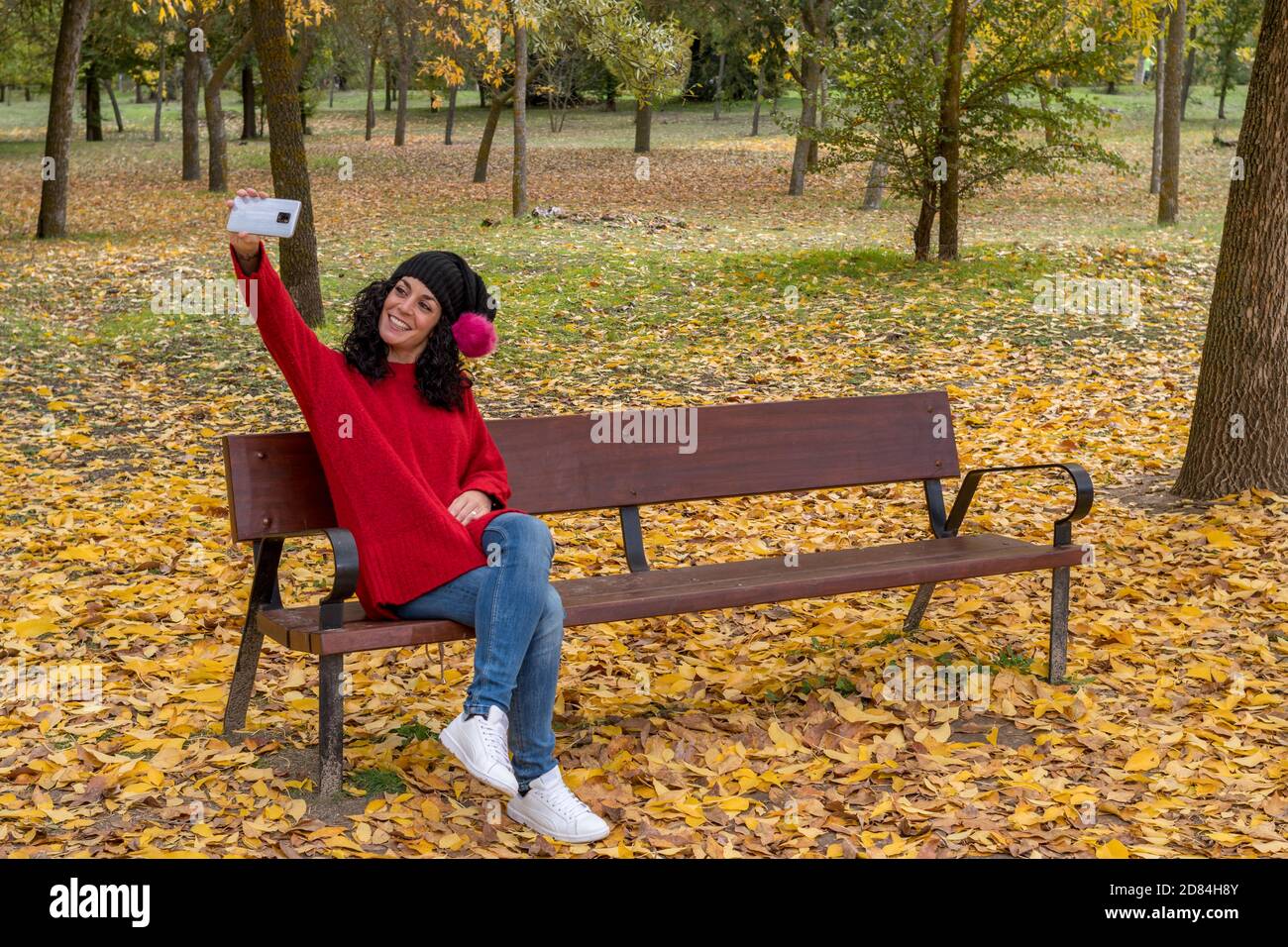 Young woman with curly black hair wearing a red woolen sweater and a black hat with a pink pom-pom sitting on a wooden bench while smiling taking a se Stock Photo