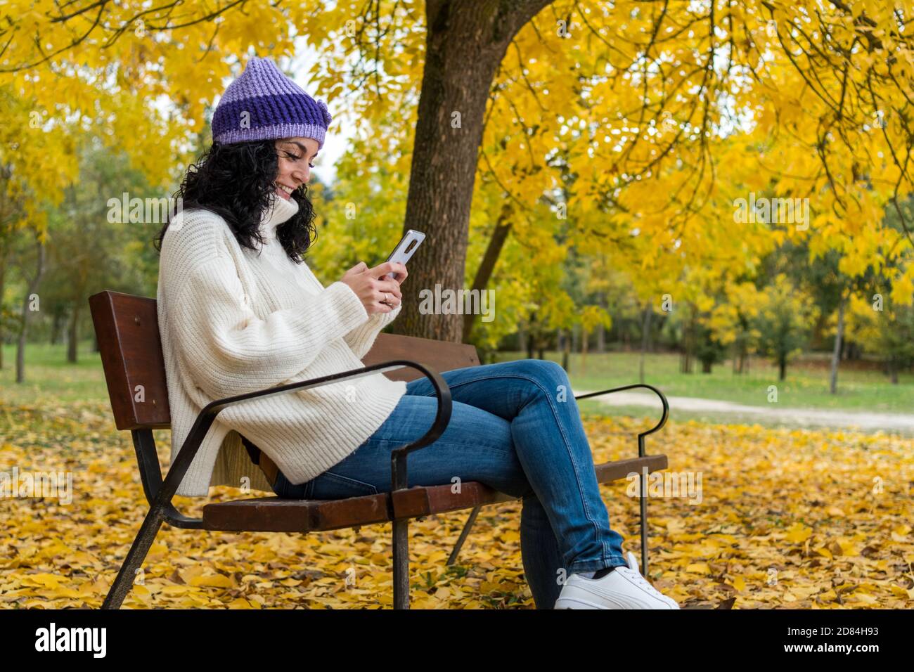 young woman with curly black hair and warm in sweater and wool hat sitting on a wooden bench in a park in autumn with leaves of the trees falling in t Stock Photo