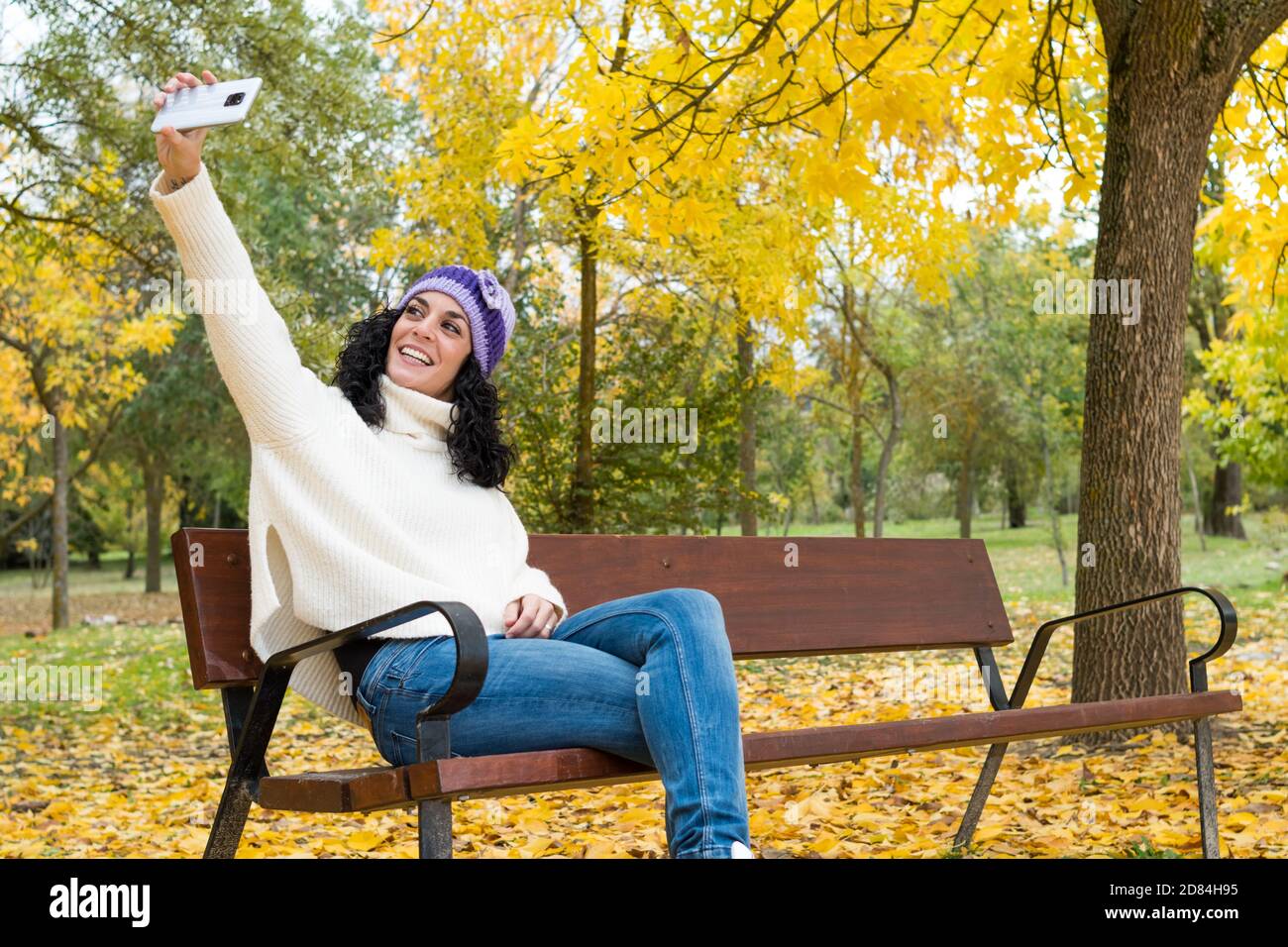 young woman with curly black hair and warm in sweater and wool hat taking a self portrait with her mobile while smiling sitting on a wooden bench in a Stock Photo