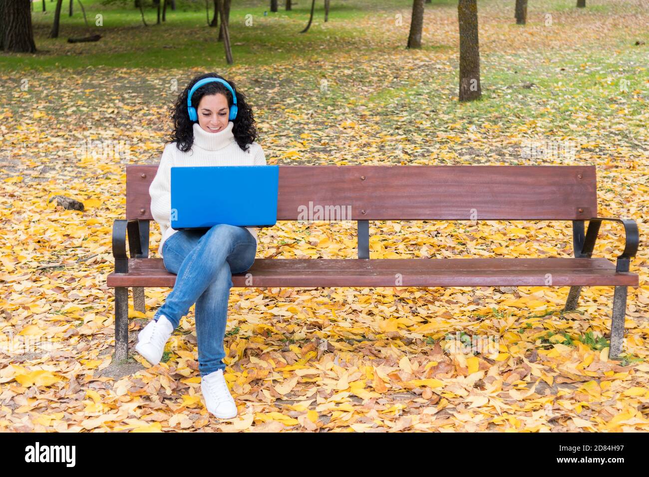 Young woman with curly black hair wearing wool sweater sitting on a wooden bench while working with her laptop and blue wireless headphones in a park Stock Photo
