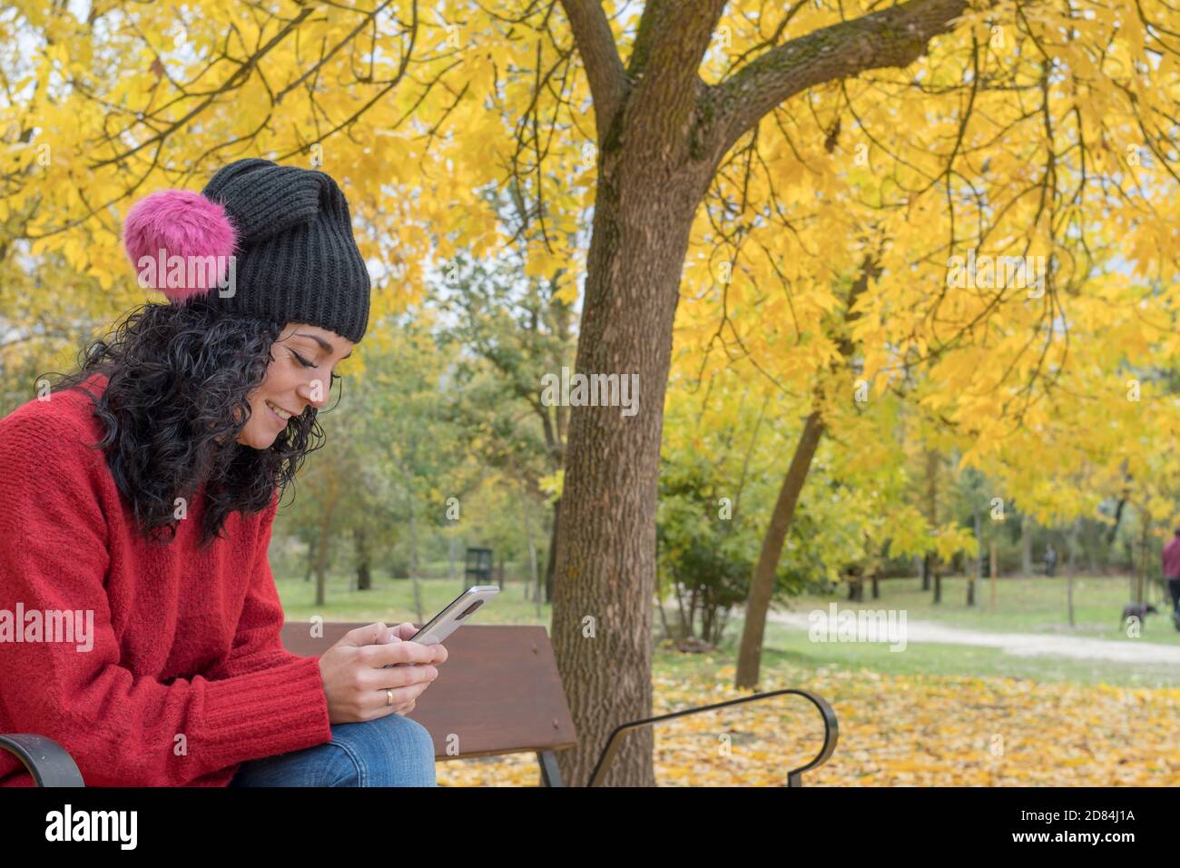 young woman with curly black hair wearing a sweater and wool hat sitting on a wooden bench in a park in autumn with leaves of the trees falling in the Stock Photo