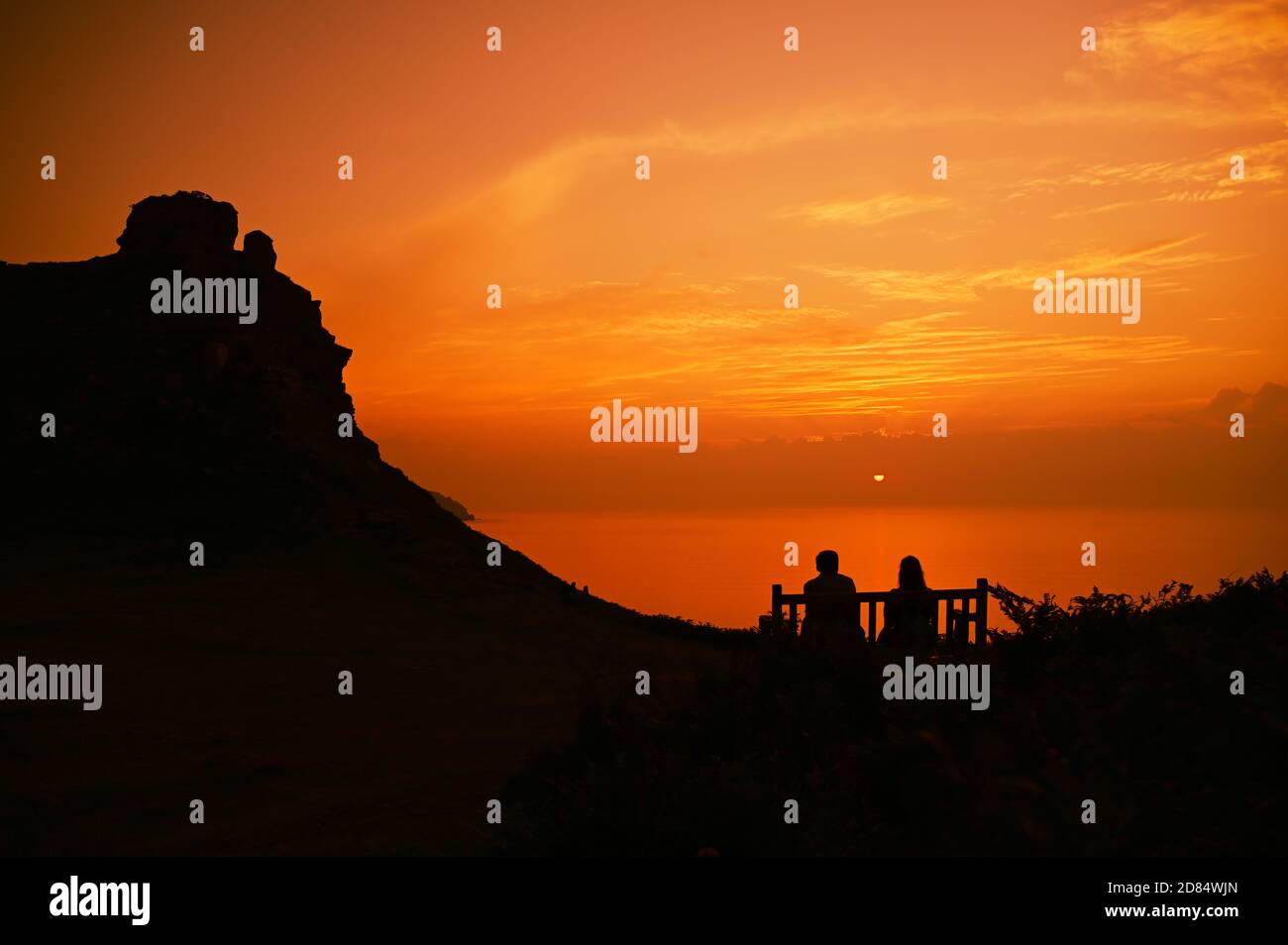 A golden sunset across the sea with a mountain on the left hand side and people sitting, silhouetted on a bench in the Valley of Rocks, Exmoor, Devon Stock Photo