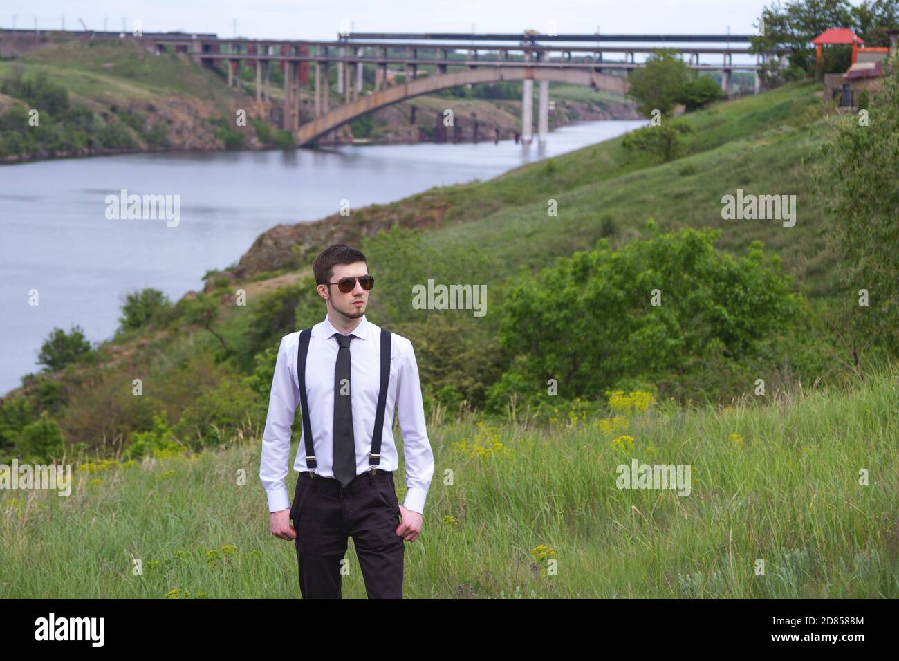 Portrait of stylish brunet young man, wearing white shirt, black tie, suspenders and glasses, walking on the meadow near river. Stock Photo