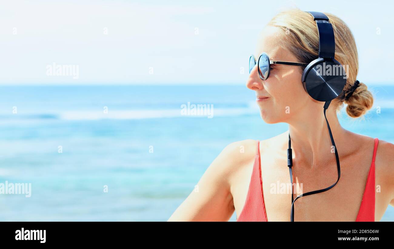 Young positive girl in headphones and sunglasses listening music and relaxing. Woman chilling at tropical sea beach. Travel family lifestyle Stock Photo