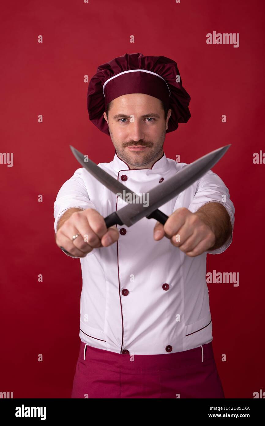 Unshaven handsome chef posing crossed knives in hands on red studio background Stock Photo