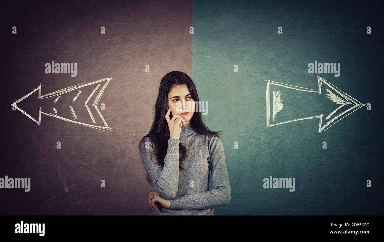 Curious and doubtful young woman, has to choose right or left side. Puzzled student girl in front of a split blackboard with arrows showing two differ Stock Photo