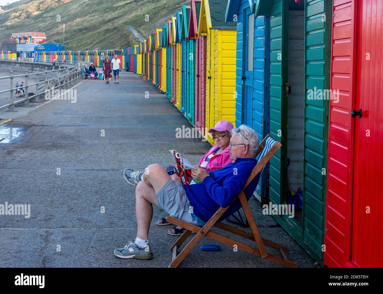 Couple sitting on deck chairs outside beach hut on promenade, Whitby