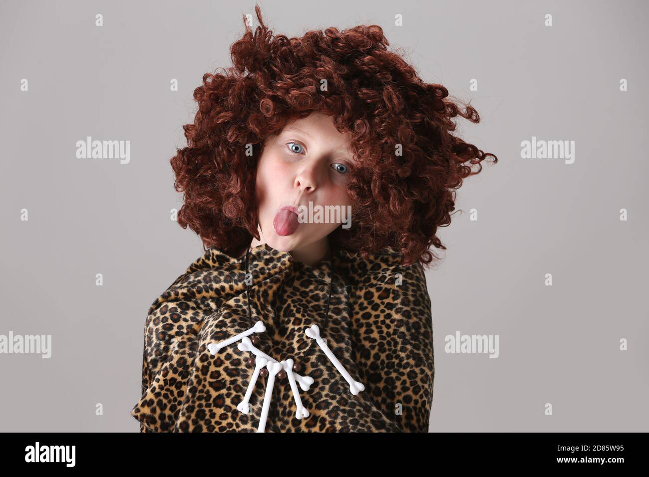 young girl in leopard skin cloak wearing  1960s, 1970s style Afro wig and a necklace made of toy bones cheekily sticking her tongue out Stock Photo