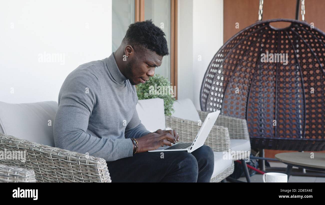 Young handsom black man working on the laptop on his balcony while drinking coffee. High quality photo Stock Photo