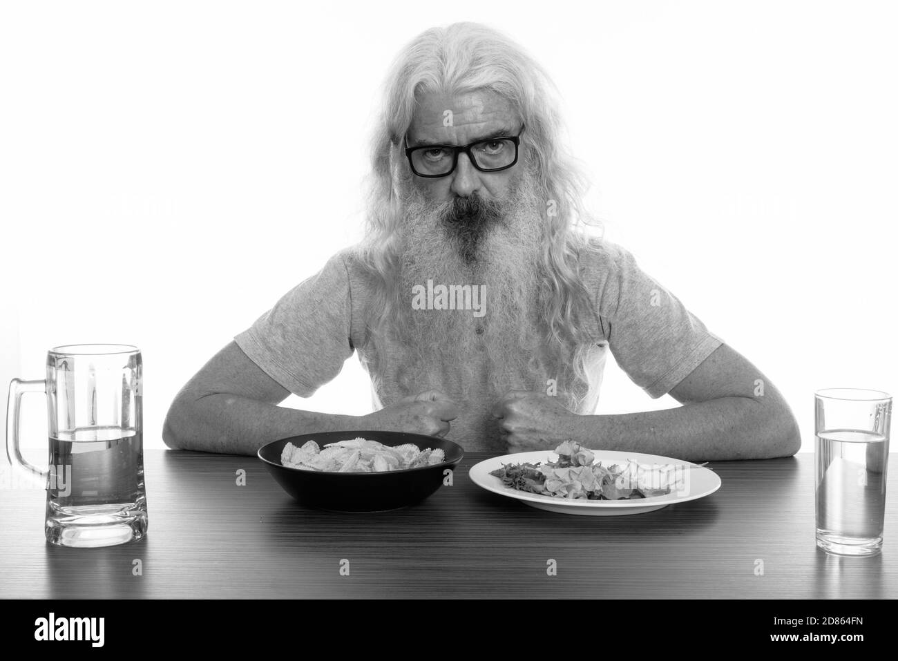 Studio shot of senior bearded man wearing eyeglasses with healthy and unhealthy foods on wooden table Stock Photo