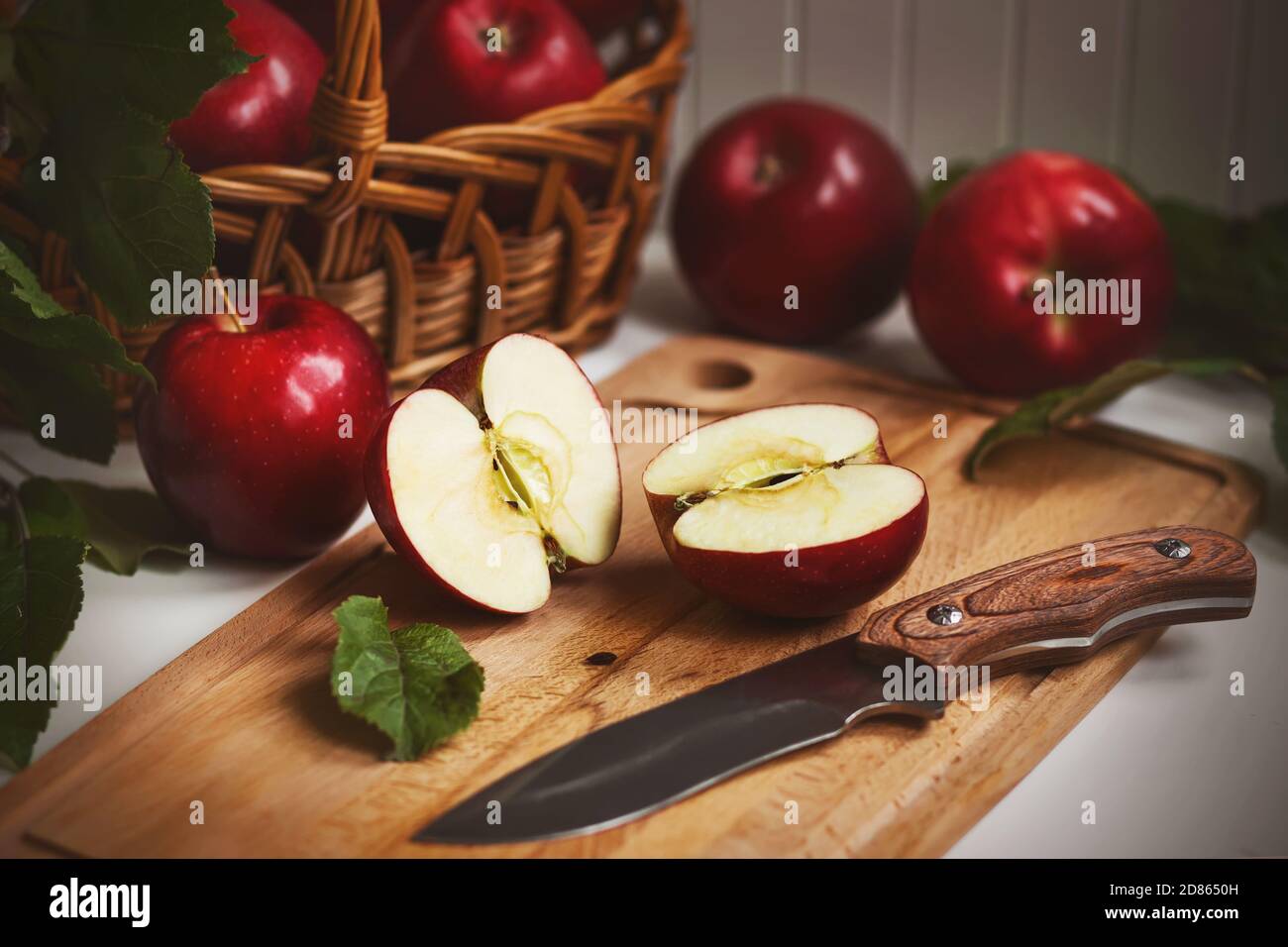 On the kitchen table still life - wicker basket with red ripe sweet apples, next to it a wooden board on which lies an Apple cut in half and a sharp k Stock Photo
