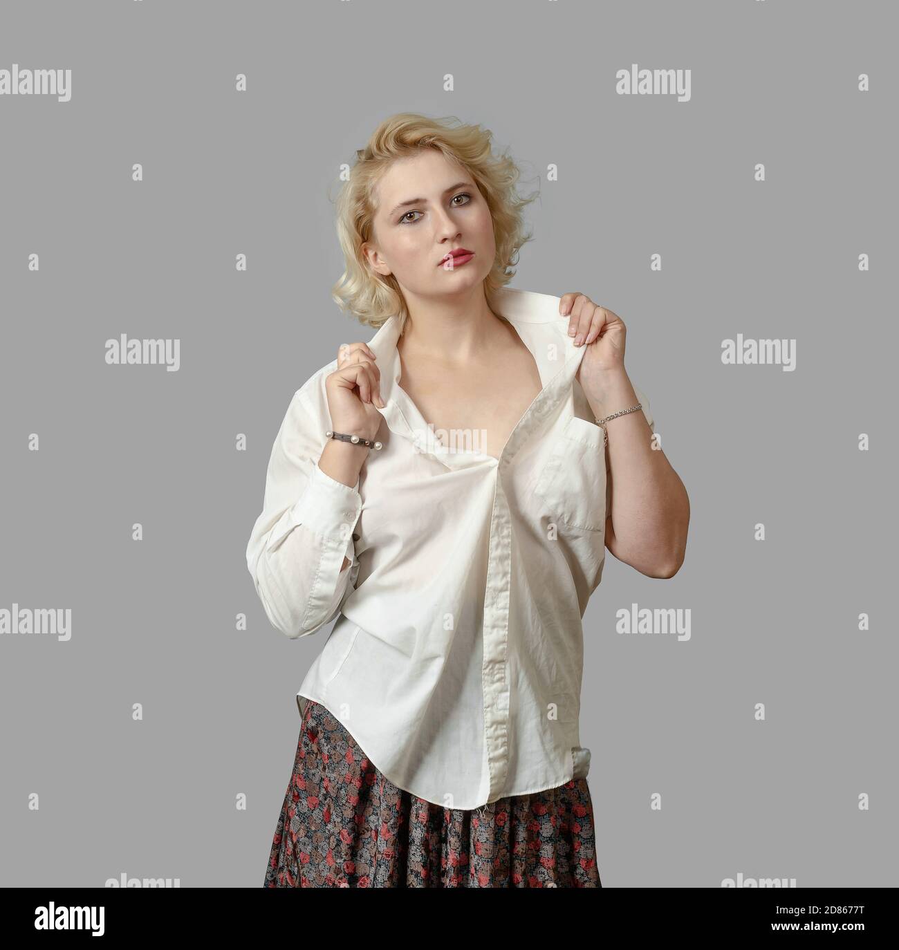 Young blonde woman in white shirt looks at camera, raises his collar on grey background. Stock Photo