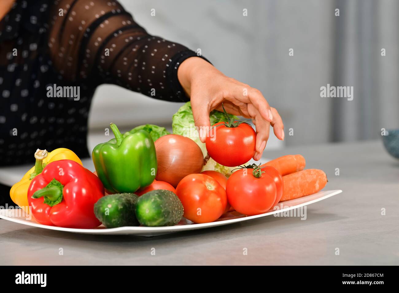 Close up of a female hand picking up a delicious looking tomato from a plate with assorted vegetables. Healthy food and vegetables concept. Stock Photo