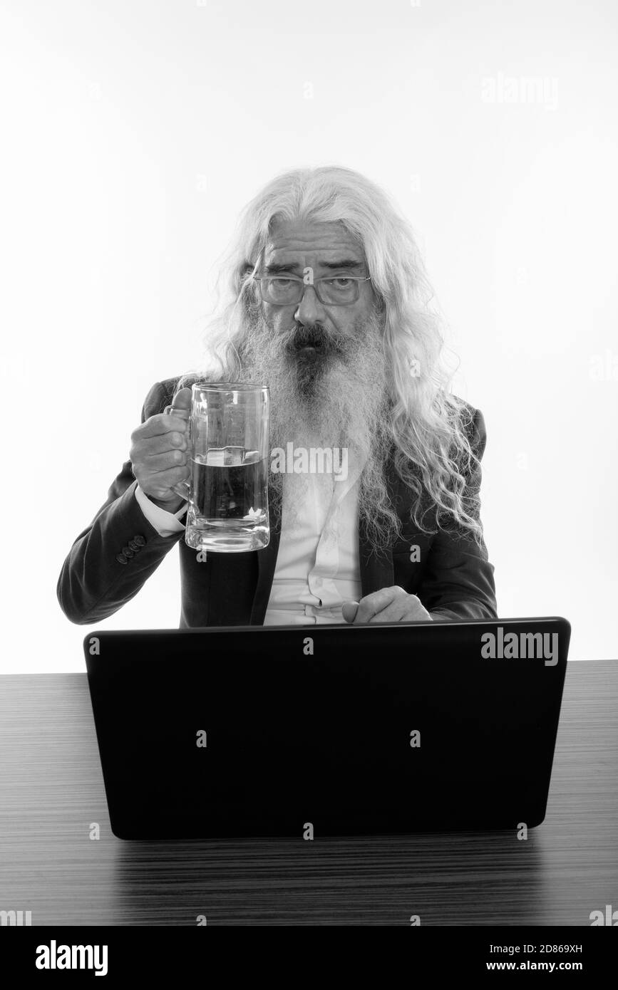 Studio shot of senior bearded businessman holding glass of beer with laptop on wooden table Stock Photo