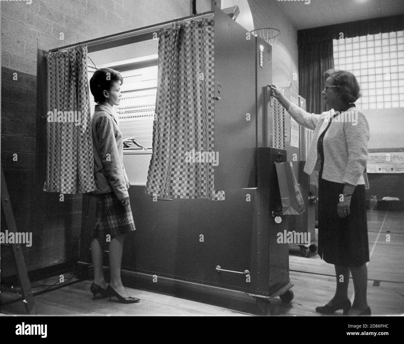 Here a voter prepares to cast her ballot by pulling a lever which closes the privacy curtain and unlocks the machine for voting. A precinct official clears the machine and registers the vote by pressing a button on a side panel, Jamestown, NY, 1965. (Photo by Automatic Voting Machine Co/RBM Vintage Images) Stock Photo