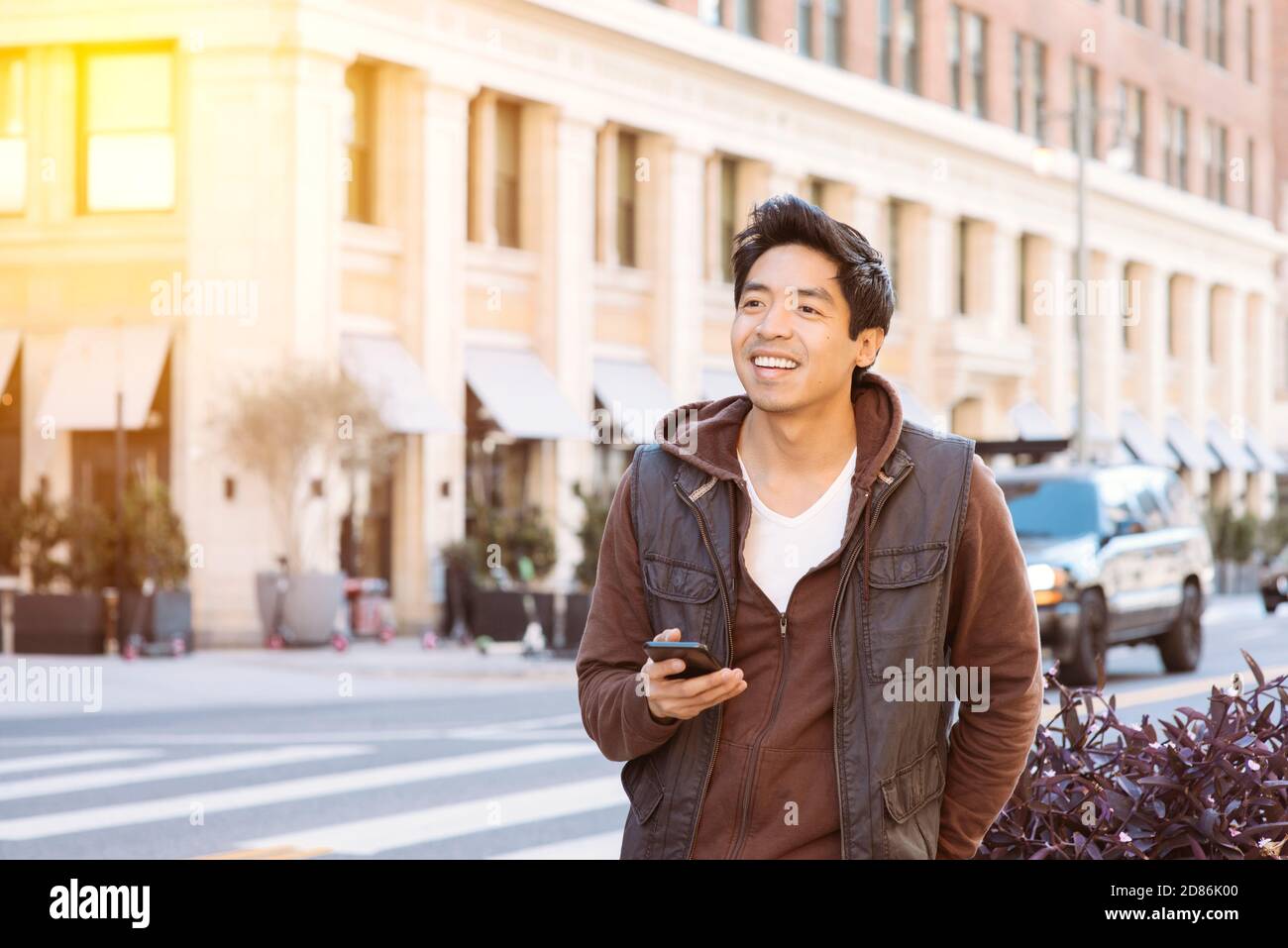 Asian male smiles and is happy waiting for his ride share while holding his smart phone - App Based transportation - City - Daytime Stock Photo