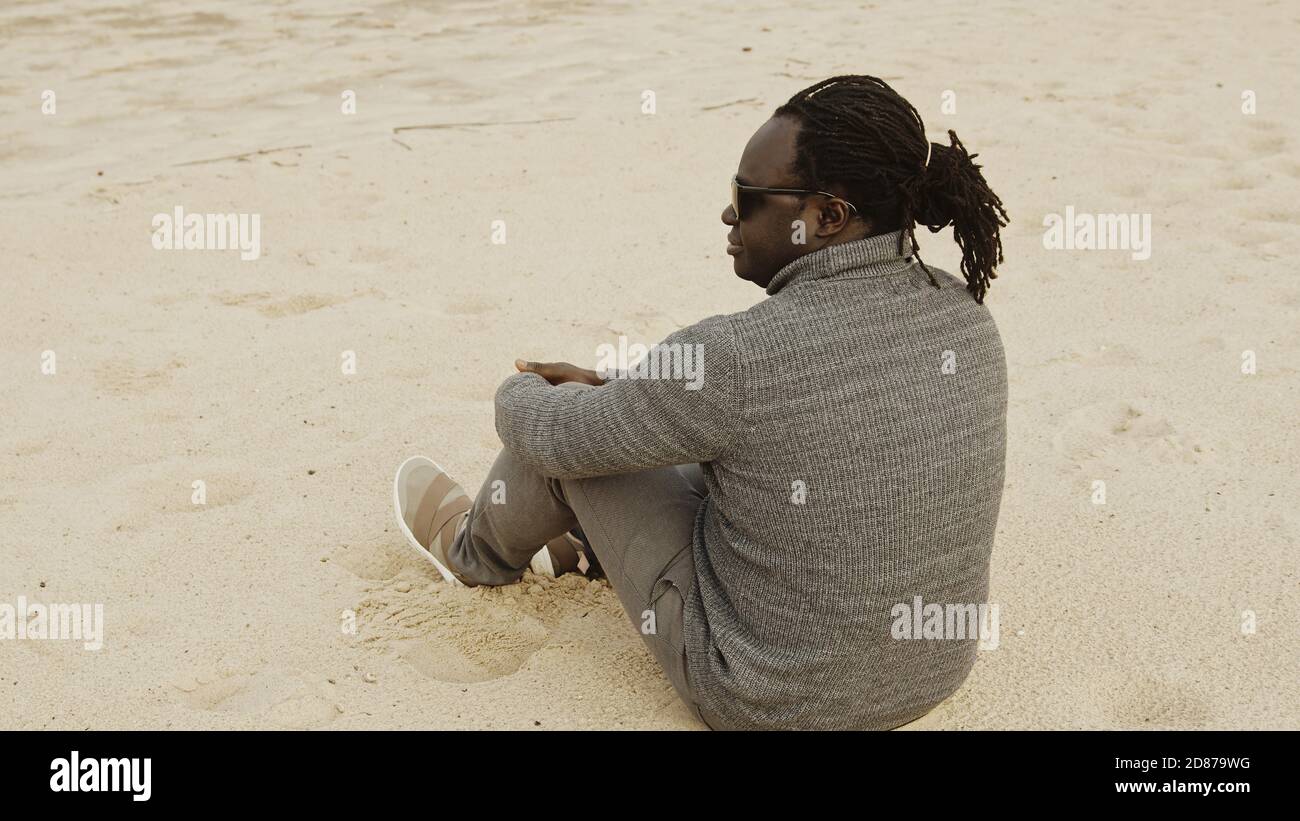 Young african man feeling cold while sitting in sweater on the sandy beach. High quality photo Stock Photo