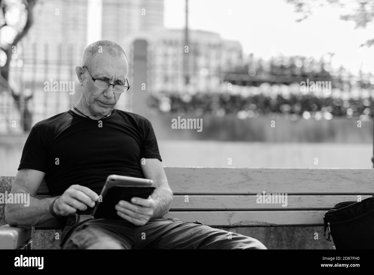 Bald senior tourist man using digital tablet while wearing eyeglasses and sitting on the wooden bench at peaceful park Stock Photo