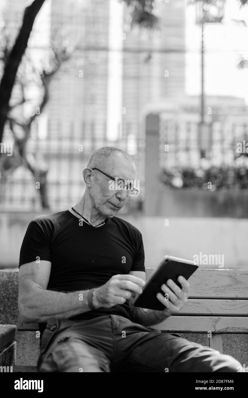 Bald senior tourist man using digital tablet on the wooden bench at peaceful park in Bangkok Thailand Stock Photo