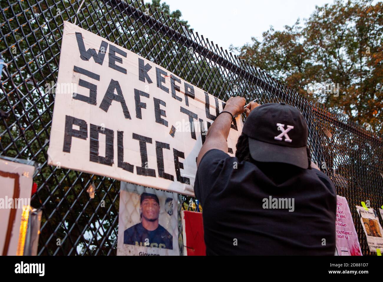 Washington, DC, USA, 27 October, 2020.  Pictured: A man zip ties a sign to the Lafayette Square fence in front of the White House.  The sign says what has become a familiar refrain in DC over the last five months, 'We keep us safe, not the police.'  The man hung the sign at an event to redecorate the fence with protest art after Trump supporters destroyed the art that was there, as MPD officers watched, on the night of 26 October, 2020. Credit: Allison Bailey/Alamy Live News Stock Photo