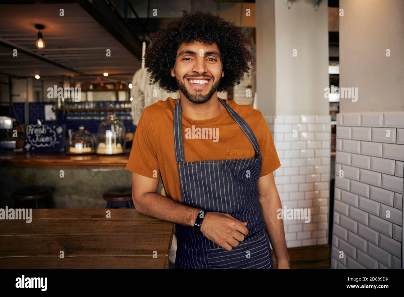 Portrait of smiling young male waiter wearing apron leaning on table standing looking at camera in cafe Stock Photo