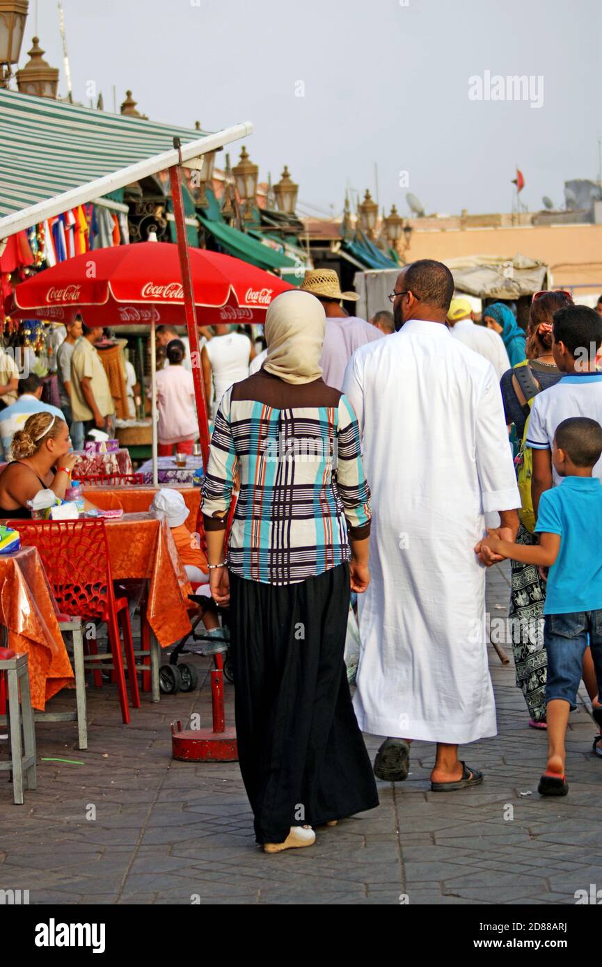 A man, woman, and child walk past a cafe in the Jemaa el-Fina in Marrakesh, Morocco.  The main square of the Medina is a UNESCO cultural space. Stock Photo