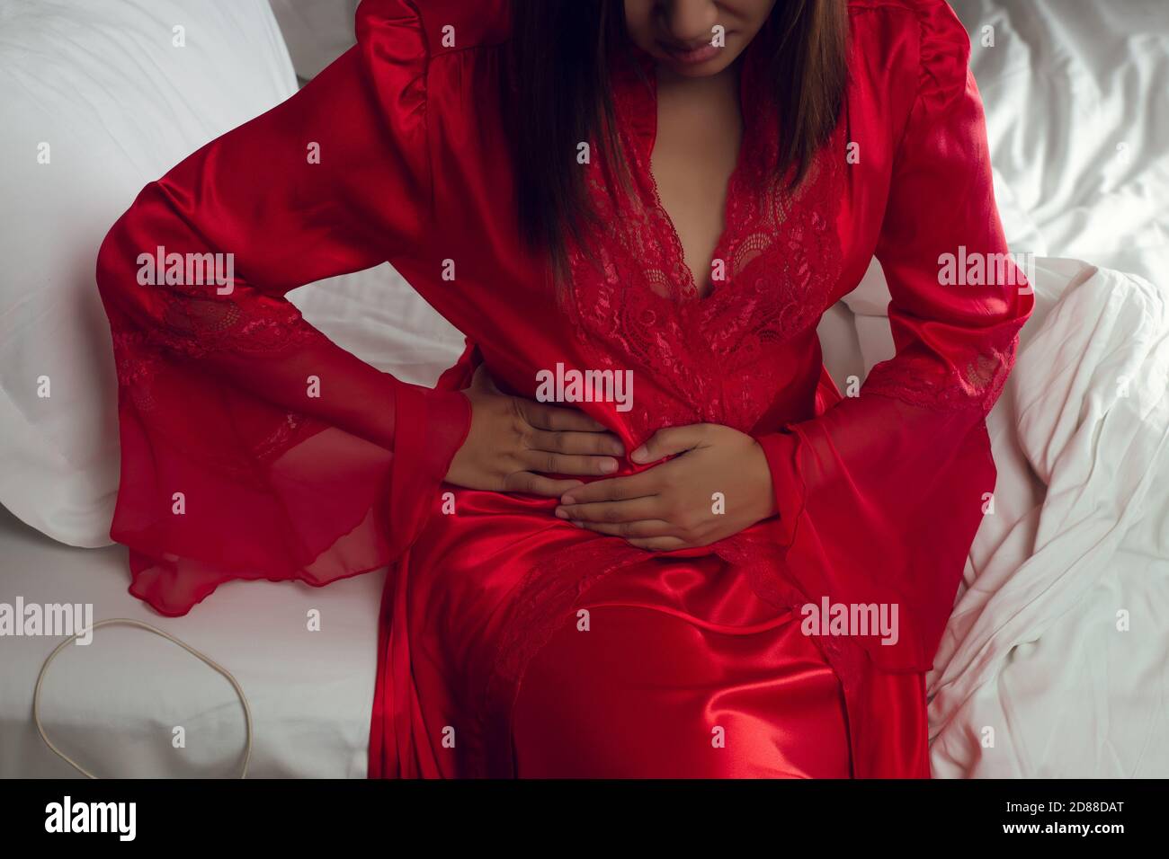 A woman in satin red nightgown suffering from menstruation pain on the bed at night. Irritable Bowel Syndrome or IBS Stock Photo