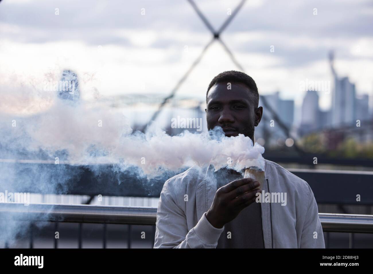 Young black man holding smoking smoke bomb with big city background. Medium  shot Stock Photo