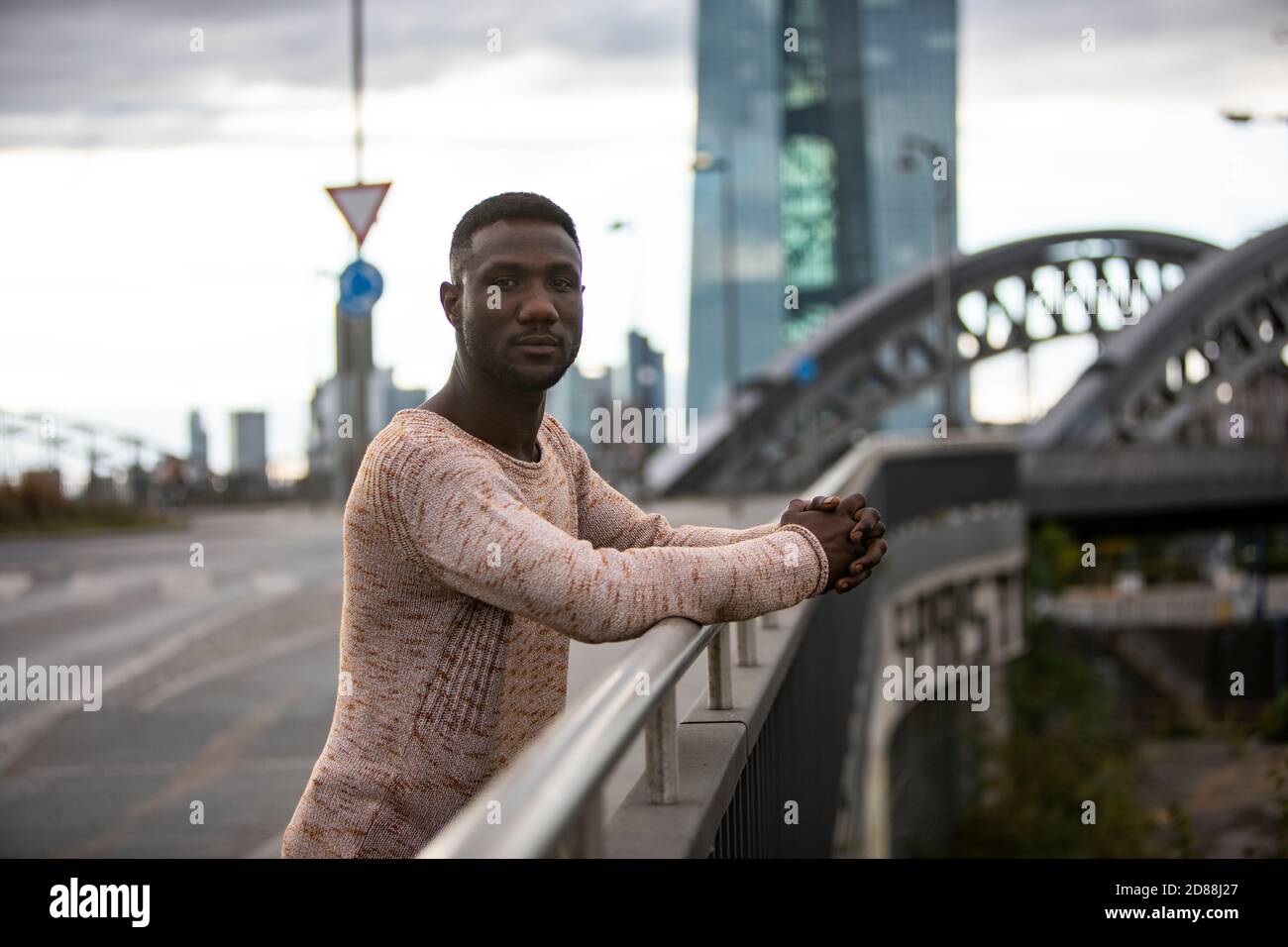 Man standing on bridge leaning arms on railing and looking at camera. Medium shot. City background. Stock Photo