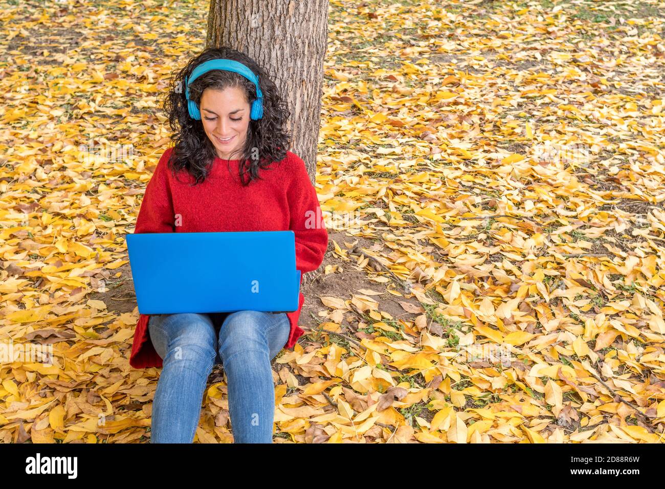 young woman with curly hair wearing red sweater sitting on the ground leaning against a tree with wireless headphones while working with her laptop in Stock Photo