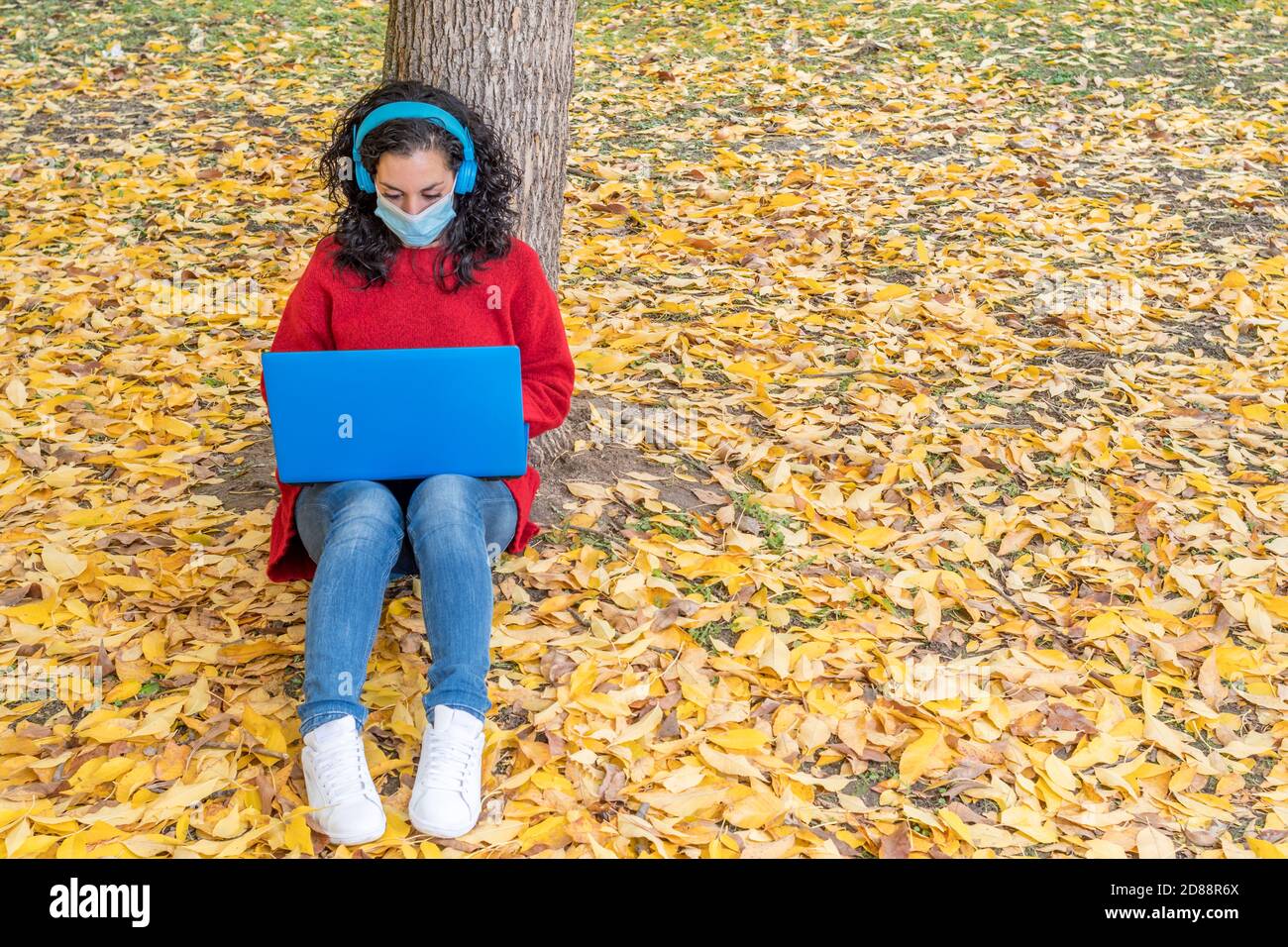 young woman with face mask due to the covid-19, coronavirus epidemic sitting on the ground with wireless headphones while working with her laptop in a Stock Photo