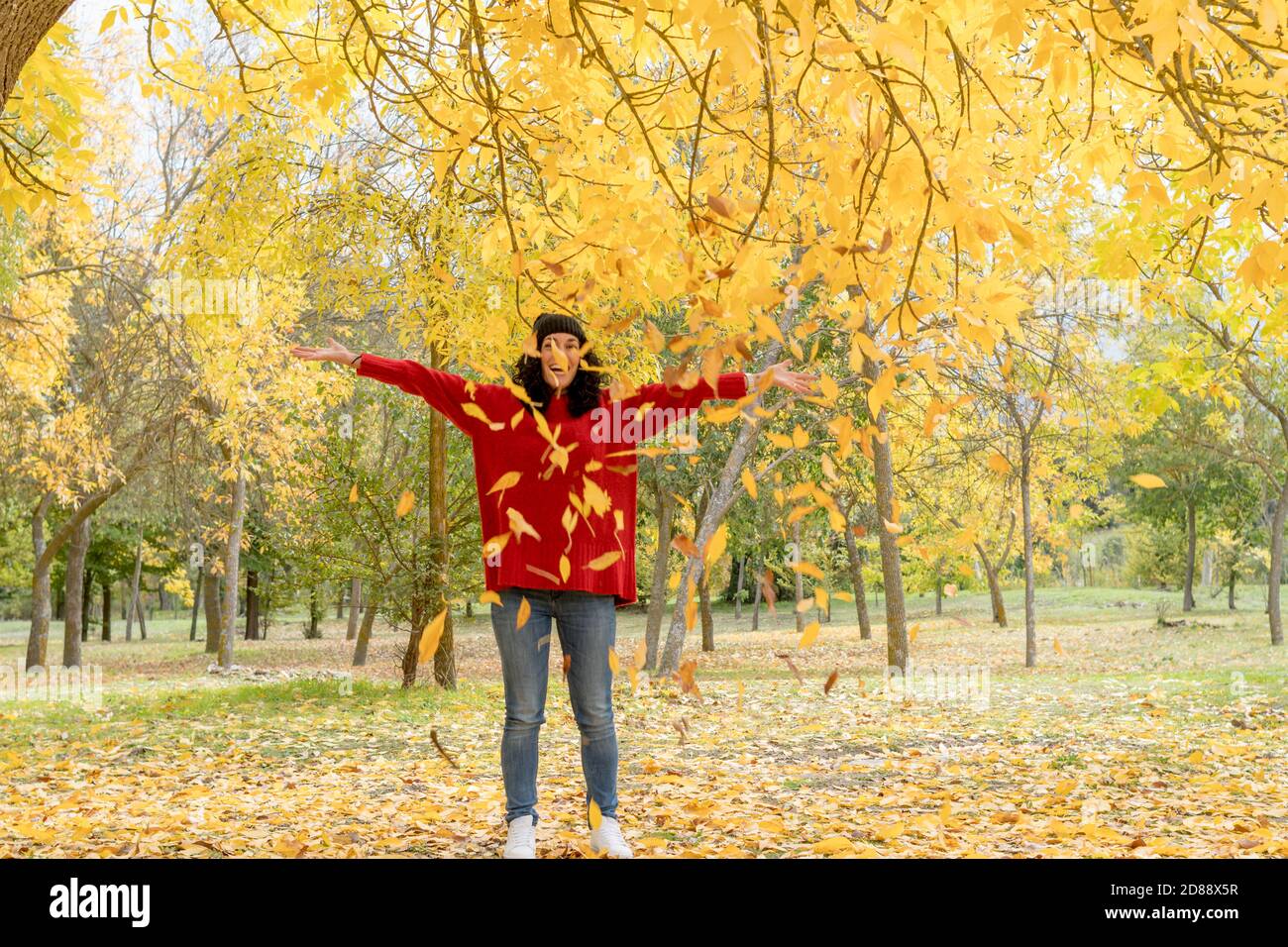 young woman with curly black hair in red sweater and woolen hat has fun throwing dry leaves that have fallen from the trees in a park in autumn Stock Photo