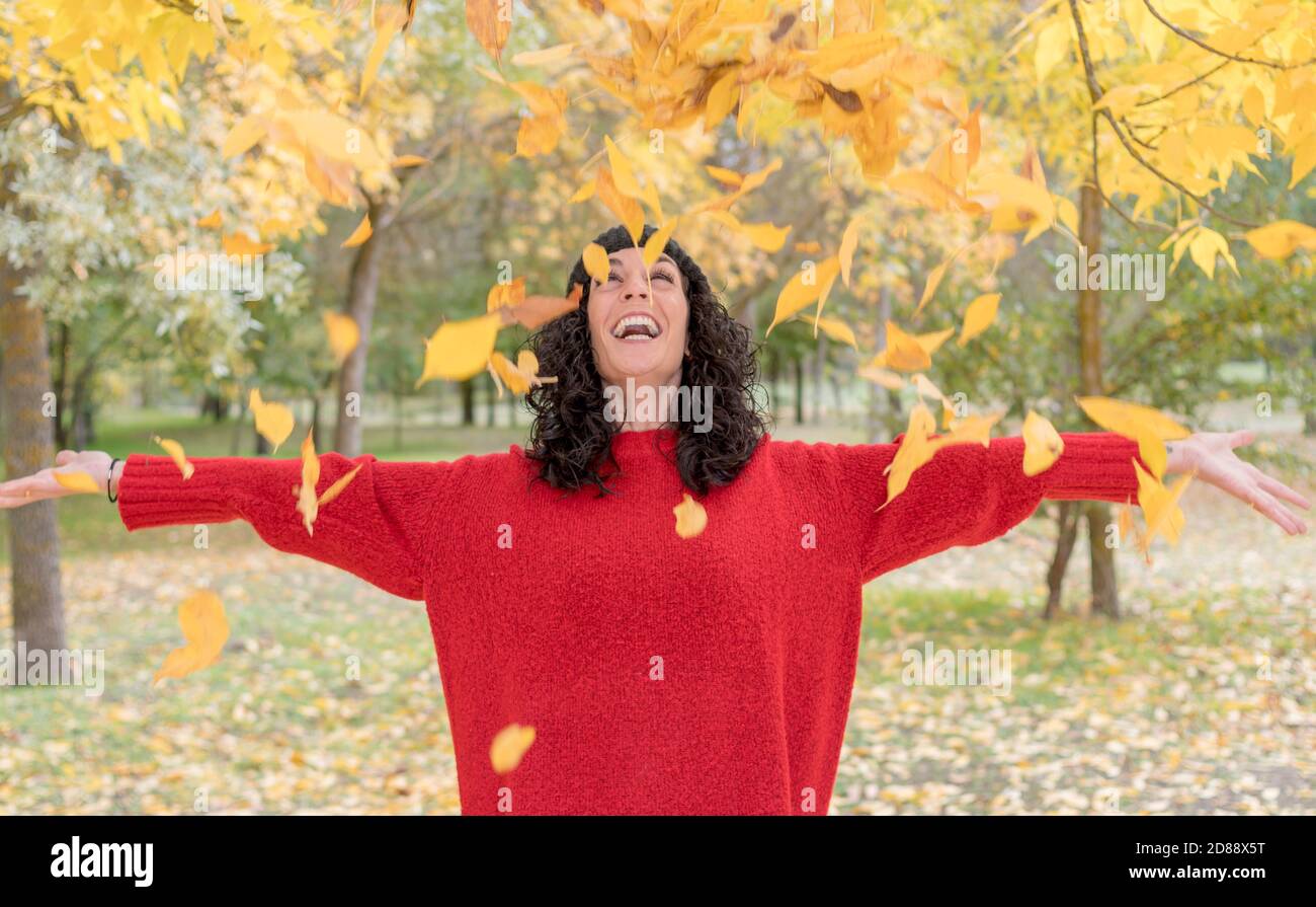 young woman with curly black hair in red sweater and woolen hat has fun throwing dry leaves that have fallen from the trees in a park in autumn Stock Photo