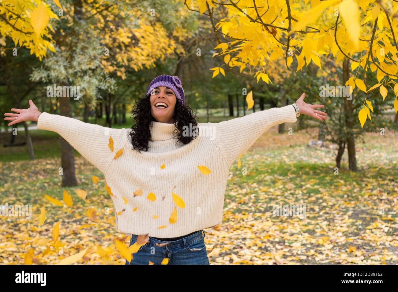 young woman with curly black hair in white sweater and knit hat has fun throwing dry leaves that have fallen from the trees in a park in autumn Stock Photo