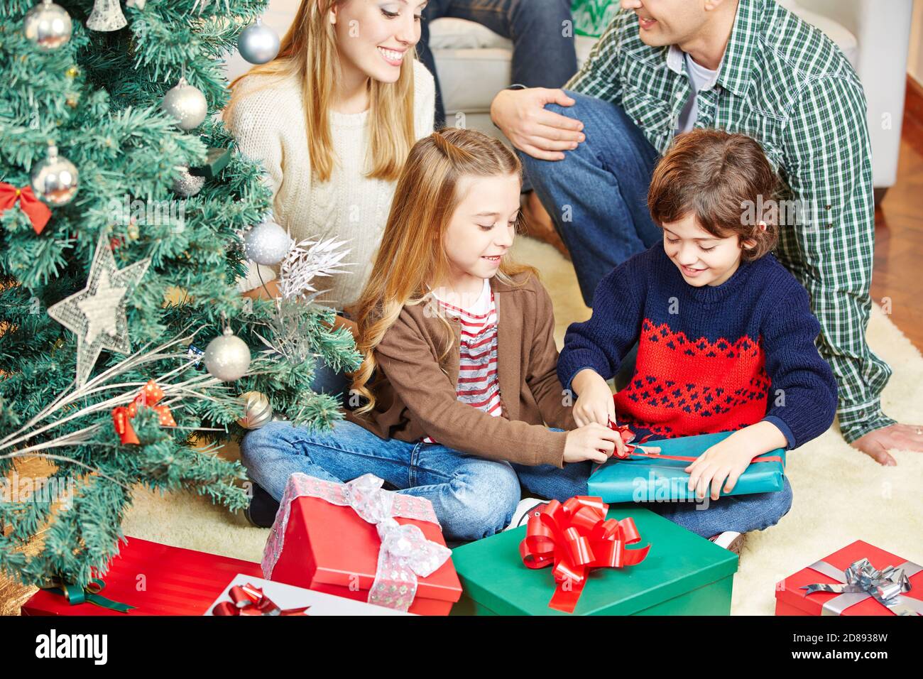 Two children open Christmas presents with family next to Christmas tree Stock Photo