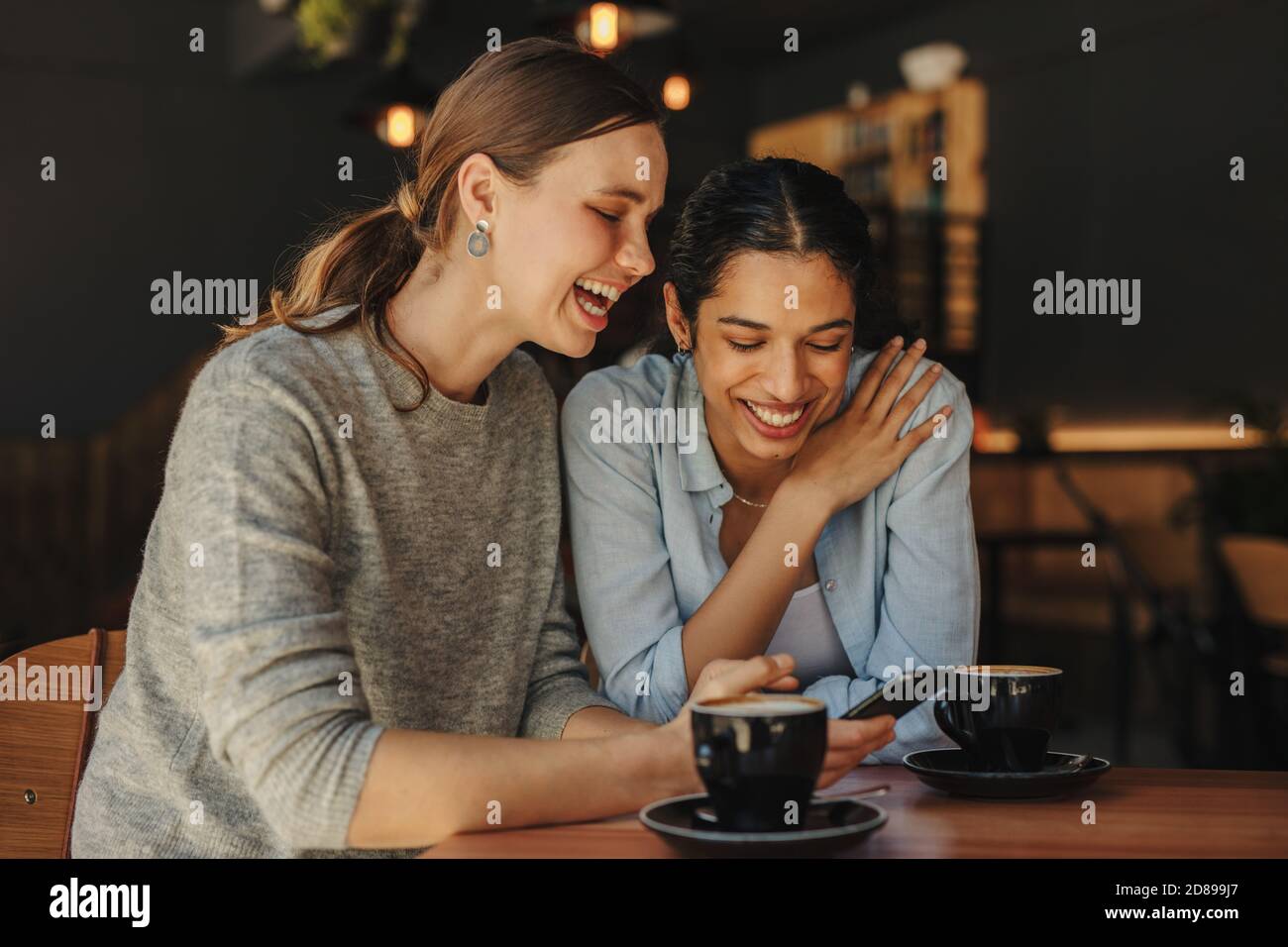 Woman showing funny posts on her cell phone to her friend and smiling. Two female friends sitting in coffeeshop using a cell phone and smiling. Stock Photo