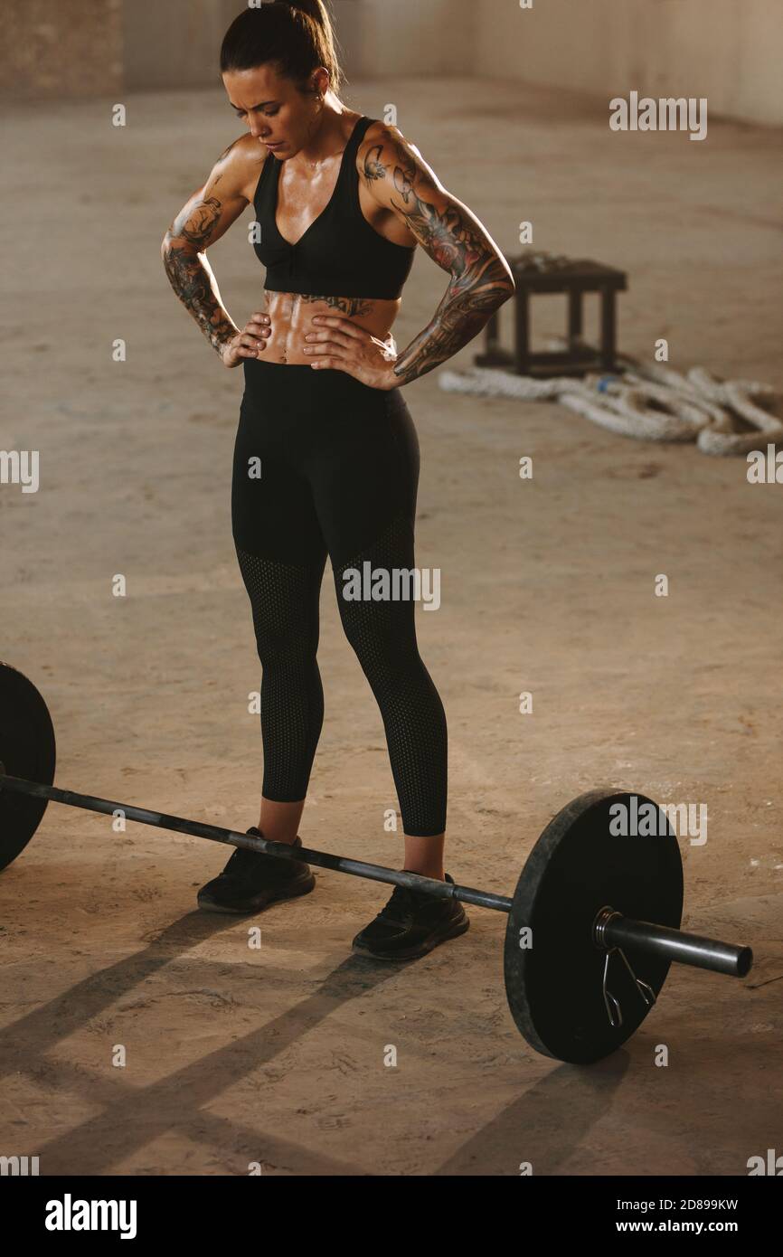 Strong woman standing with her hands on hips at cross training space with barbell in floor. woman in sportswear about to start workout with heavy weig Stock Photo