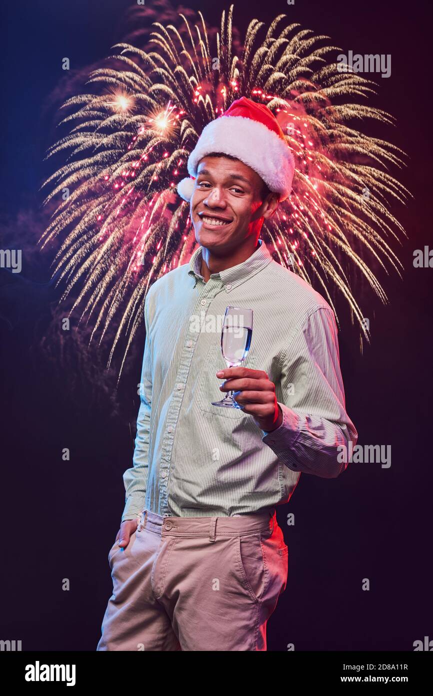 Happy New Year. Vertical shot of a young cheerful mixed race man wearing Santa Claus cap with glass of champagne standing against fireworks background and smiling. Christmas party, people and holidays Stock Photo